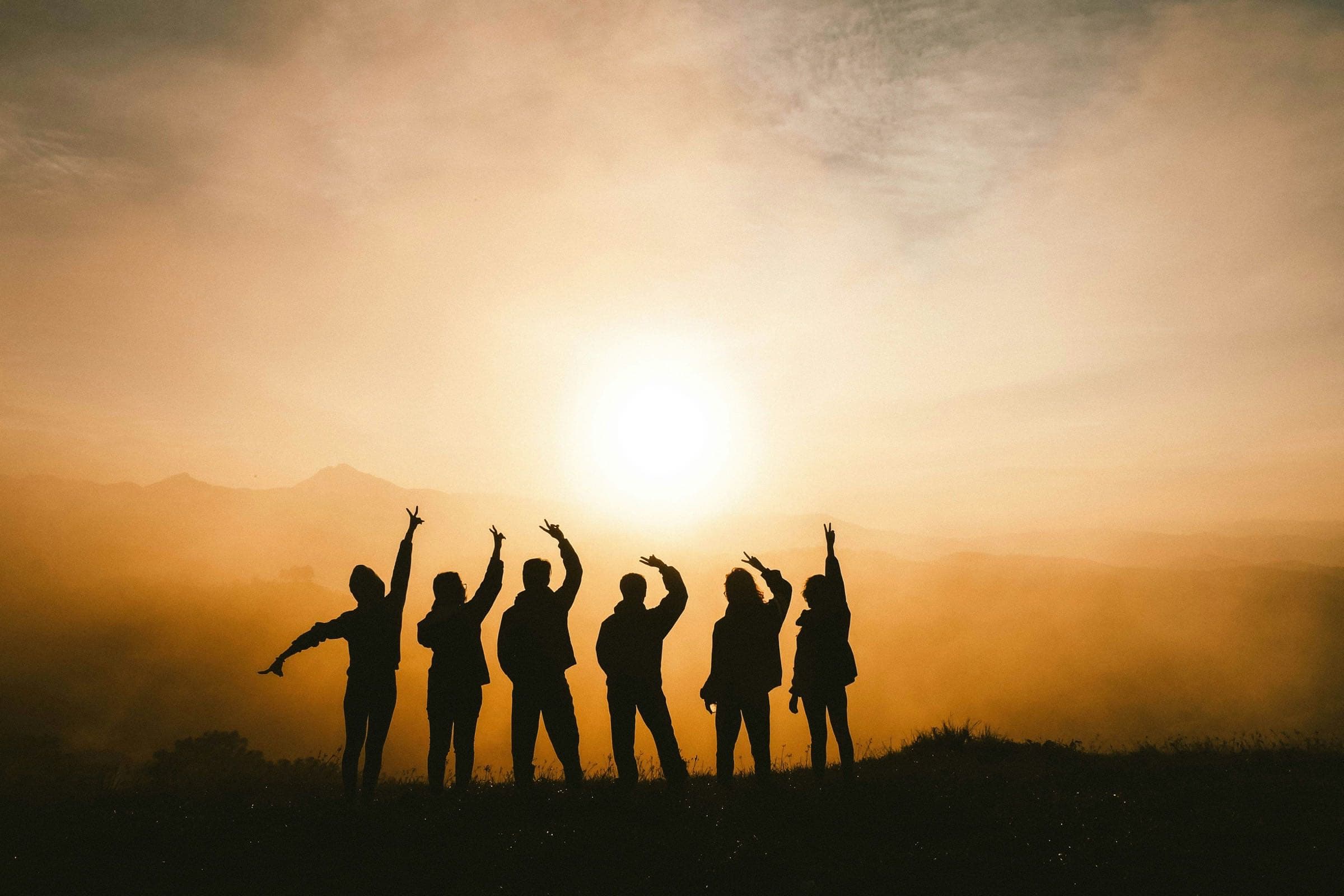 Seis amigos em silhueta no cimo de uma colina ao nascer do sol, de braços erguidos para o sol
