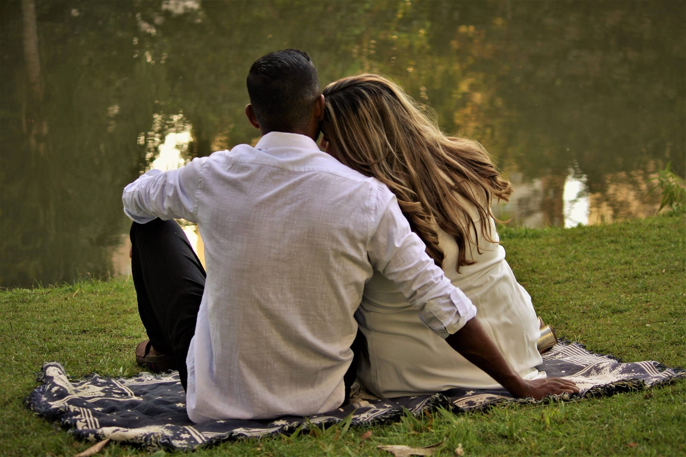 Um casal sentado lado a lado numa manta junto a um lago, visto de costas